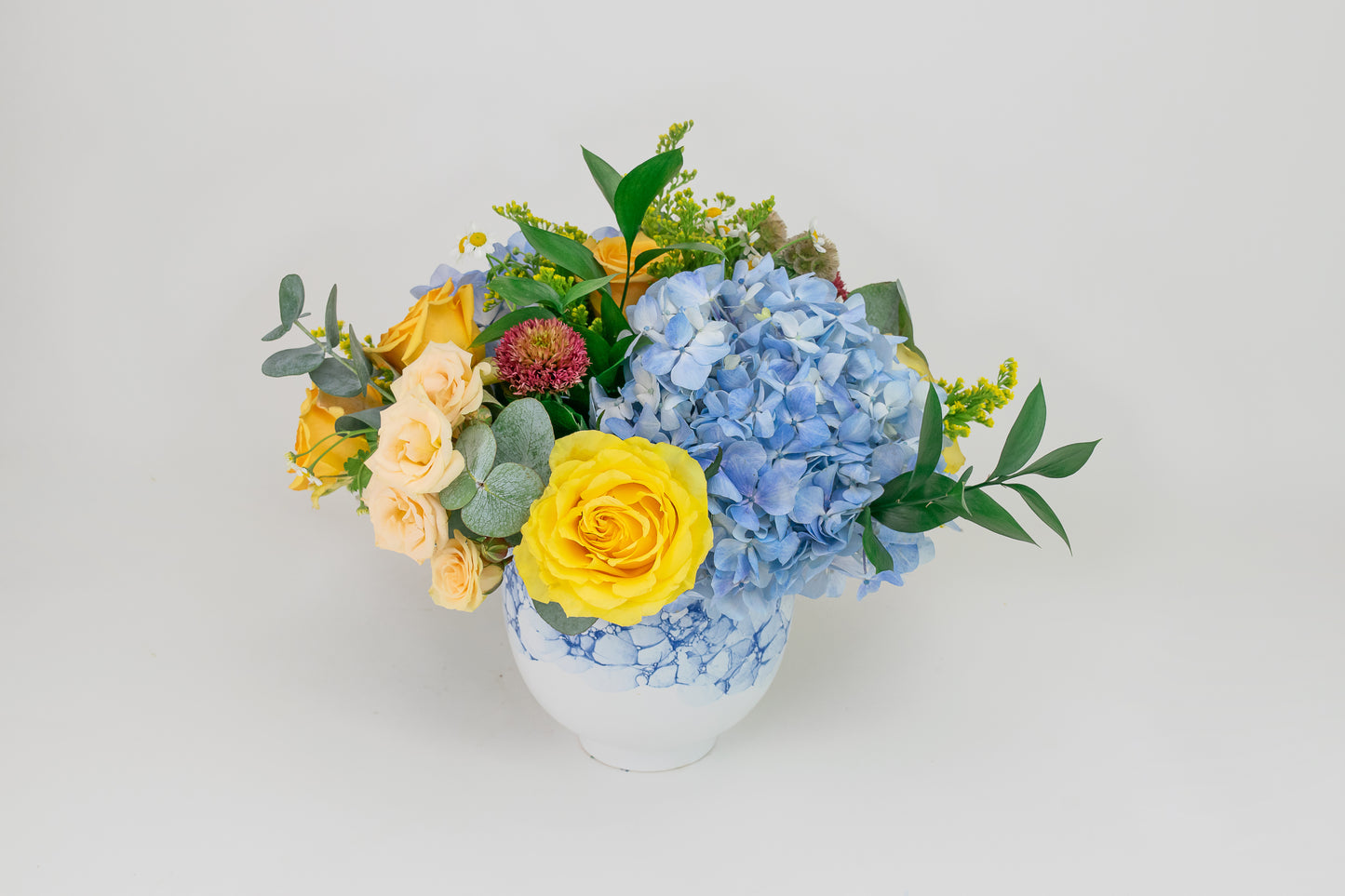 Floral arrangement with yellow roses and blue hydrangeas in a white vase on a light gray background