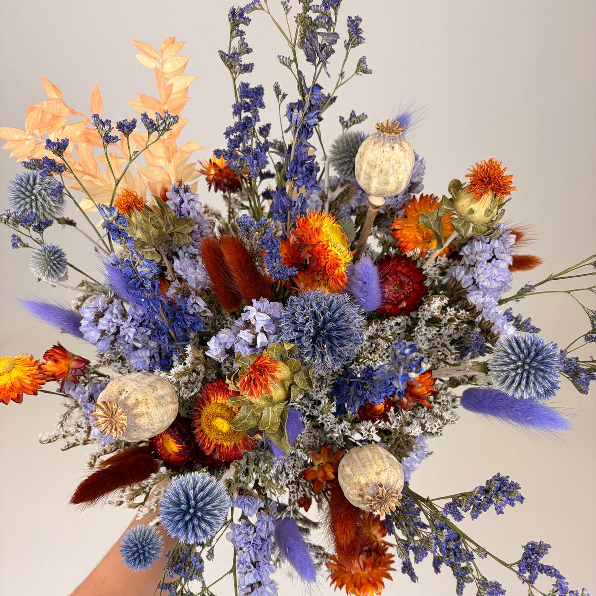 A dried flower bouquet held by a person's hand against a plain light gray background. The bouquet features a mix of blue, orange, purple, beige, and brown dried flowers and botanicals.