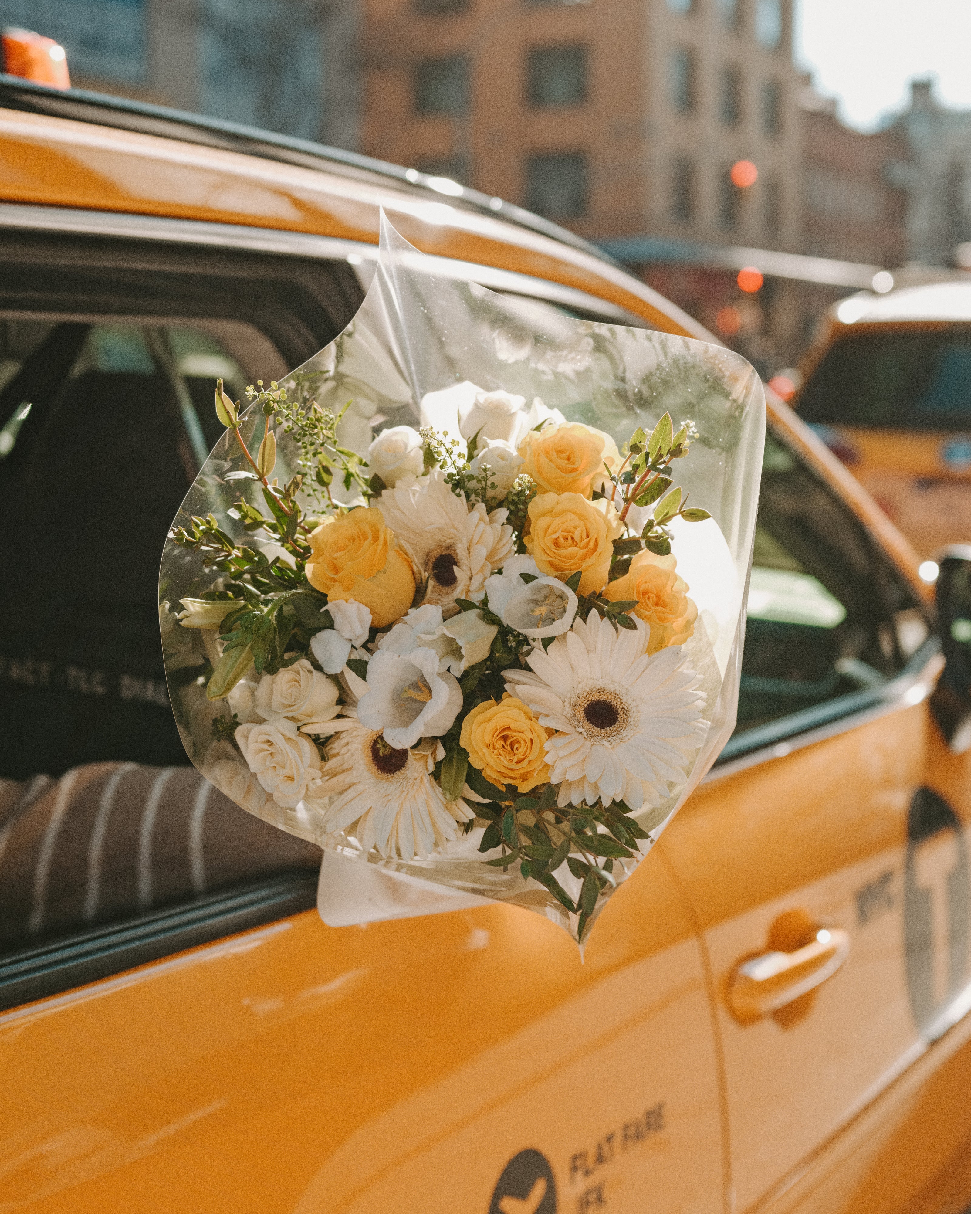 Bouquet of yellow and white flowers in clear plastic wrap held by a hand reaching out of a yellow taxi cab.