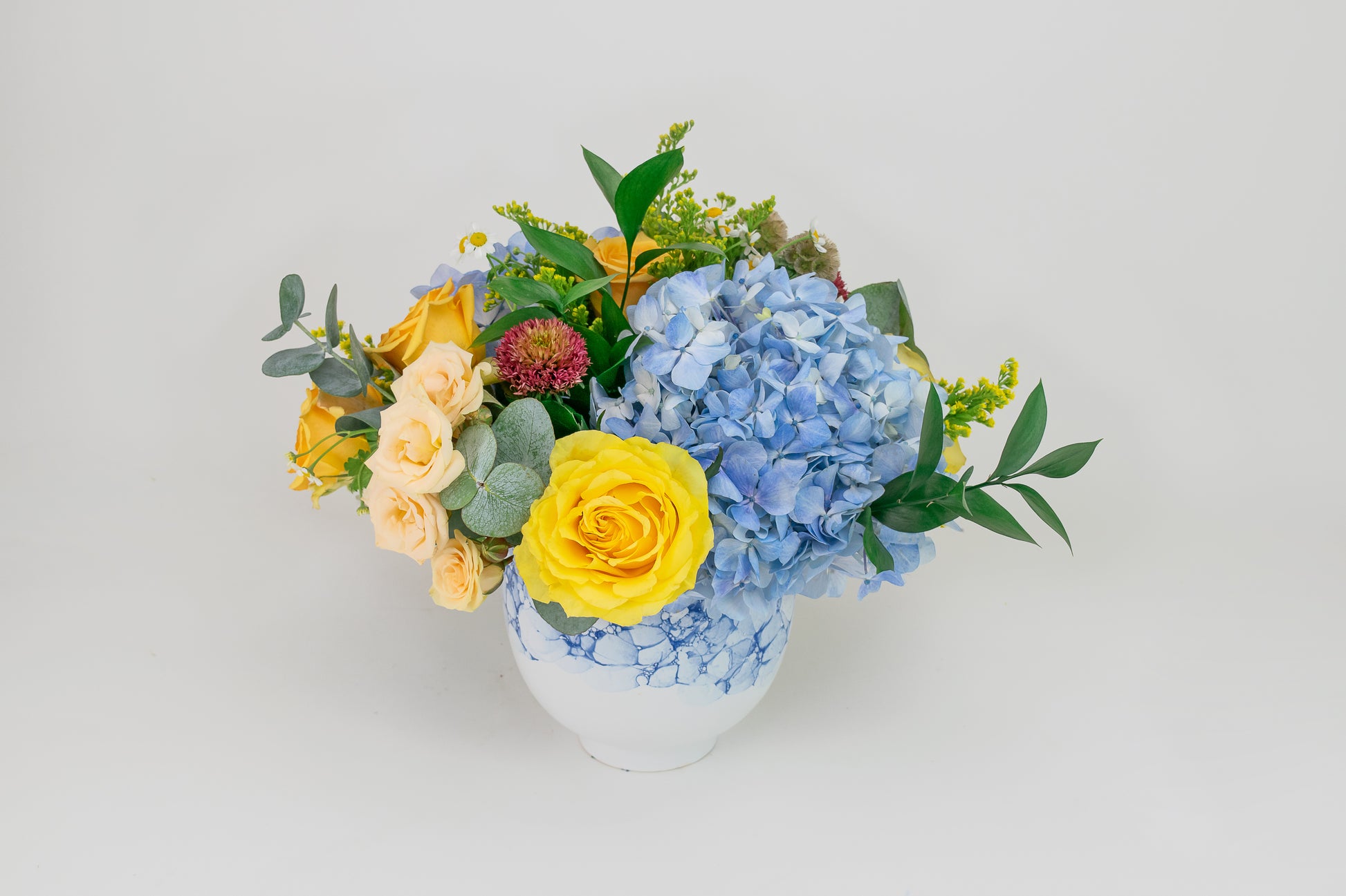 Floral arrangement with yellow roses and blue hydrangeas in a white vase on a light gray background